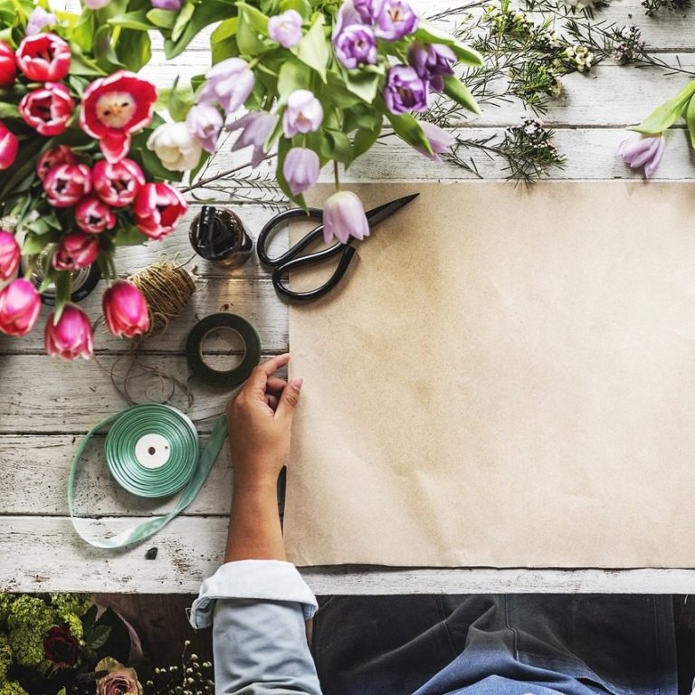 Mesa con flores, papel kraft y herramientas de jardinería. Mano sostiene tijeras.